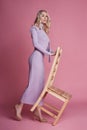 Fashion Beautiful blonde woman in knitted closed long dress stands near the chair on a pink background. Perfect hair and makeup Royalty Free Stock Photo