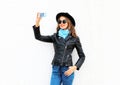 Fashion cool young smiling girl taking picture self portrait on smartphone in city wearing black rock jacket, hat over white Royalty Free Stock Photo