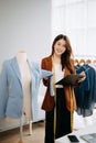 Fashion designer woman talking smart phone and using laptop with digital tablet computer in modern studio the clothes hanging on Royalty Free Stock Photo