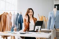 Fashion designer woman talking smart phone and using laptop with digital tablet computer in modern studio the clothes hanging on Royalty Free Stock Photo