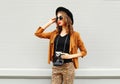 Fashion look, pretty cool young woman model with retro film camera wearing elegant hat, brown jacket outdoors over city background Royalty Free Stock Photo