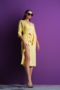 Fashion model in big sunglasses wearing yellow dress with unfastened buttons near purple wall