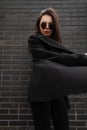 Fashion model comely young stylish woman in trendy sunglasses in jacket in jeans posing near vintage brick wall in city. Cool fine Royalty Free Stock Photo