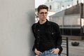 Fashion model handsome young man in fashionable denim clothes in glasses with backpack resting near wall in city. Urban hipster Royalty Free Stock Photo