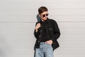 Fashion model handsome young man with trendy hairstyle in sunglasses in black denim jacket in blue jeans with backpack posing near