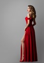 Fashion Model in Long Red Dress. Elegant Woman in evening Gown over Gray. Sexy Girl showing Leg in Slit. Women Wavy Curly Royalty Free Stock Photo