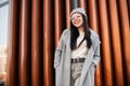 Fashion model positive young woman with cute smile in fashionable hat in glasses in long spring coat posing near modern metal wall Royalty Free Stock Photo