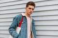 Fashion model pretty glamorous young man in stylish blue denim jacket with trendy fabric shopper rest near white wall in city. Royalty Free Stock Photo