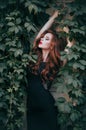 Fashion model with red hair in a long black dress posing against a wall with green leaves. A white-skinned redhead with