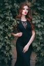 Fashion model with red hair in a long black dress posing against a wall with green leaves. A white-skinned redhead with