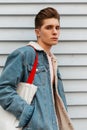 Fashion model serious glamorous young man in blue denim stylish jacket with trendy fabric shopper resting near white wall in city