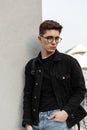 Fashion model serious young man in stylish jeans youth casual wear in fashionable glasses with backpack rest near wall in city.