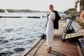 Fashion model strikes a pose on a pier near water, adorned in luxurious attire long white dress and a black blazer Royalty Free Stock Photo