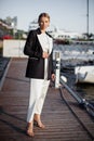Fashion model strikes a pose on a pier near water, adorned in luxurious attire long white dress and a black blazer Royalty Free Stock Photo