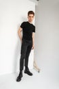 Fashion model stylish young man in vintage black t-shirt in fashionable gray youth jeans posing near white wall in room. Royalty Free Stock Photo