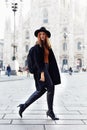 Fashion model walking by the Duomo of Milan looking back wearing a hat and a dark coat and a backpack