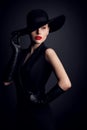 Fashion Model in Wide Brim Hat with Red Lips Make up. Elegant Retro Woman in Black Suit Dress and Gloves over Dark Gray Studio Royalty Free Stock Photo