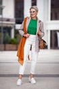 Fashion model 50 years stands street city full length, wears stylish colorful outfit, T-shirt, jacket, bag, white pants