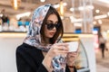 Fashion model young beautiful woman in stylish silk shawl in trendy sunglasses in black fashionable coat with cup of coffee is Royalty Free Stock Photo