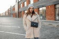 Fashion model of a young woman in fashionable coat in a knitted stylish hat with a trendy leather black handbag posing near a Royalty Free Stock Photo