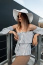 Fashion model young woman in round stylish sunglasses in white trendy straw hat in elegant striped dress posing on street. Cool Royalty Free Stock Photo