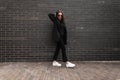 Fashion model young woman in trendy sunglasses in stylish casual youth clothes in white sneakers posing near vintage black brick Royalty Free Stock Photo