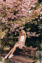 Beautiful sensual woman with blond hair in elegant clothes posing in garden with flowering sakura trees