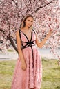 Sensual woman with blond hair in elegant dress and accessories posing among blossoming peach trees in park Royalty Free Stock Photo