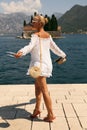 Beautiful sensual woman with blond hair in elegant dress and straw hat and bag, posing in Montenego`s city Perast Royalty Free Stock Photo