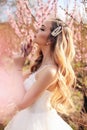 Beautiful girl with blond hair in elegant wedding dress posing among flowering peach trees in garden