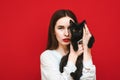 Fashion photo of attractive girl with red lips and cat on red background, looking into camera with serious face. Model poses on