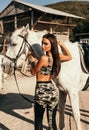 Beautiful woman with dark hair in elegant sportive suit posing near white horse in stable Royalty Free Stock Photo