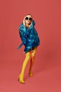 Alluring skinny young blonde woman posing in studio on red background. She is wearing blue down jacket, sunglasses Royalty Free Stock Photo