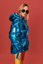 Alluring skinny young blonde woman posing in studio on red background. She is wearing blue down jacket, sunglasses Royalty Free Stock Photo