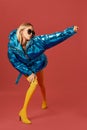 Alluring skinny young blonde woman posing in studio on red background. She is wearing blue down jacket, sunglasses Royalty Free Stock Photo