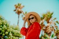 Fashion photo of a girl in black glasses with a chain and a large straw hat, a red dress on a background of palm trees. Wind in Royalty Free Stock Photo