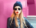Fashion portrait blonde woman sending sweet air kiss in rock black style jacket, hat posing on city street over colorful pink wall Royalty Free Stock Photo