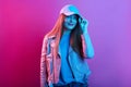 Fashion portrait of confident woman standing against pink neon wall and touching visor of her cap, wearing leather jacket, looking Royalty Free Stock Photo