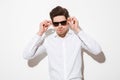 Fashion portrait of fancy man model dressed in shirt touching sunglasses and looking on camera with serious gaze, over white back Royalty Free Stock Photo