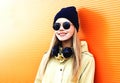 Fashion portrait happy blonde woman with headphones, sunglasses and black hat on orange colorful background in city Royalty Free Stock Photo