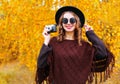 Fashion portrait pretty smiling woman with retro camera wearing autumn black hat sunglasses and knitted poncho over sunny yellow Royalty Free Stock Photo