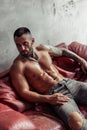 Fashion portrait of naked male model with tattoo and a black beard sitting in hot pose on red leather sofa. Loft room interio Royalty Free Stock Photo