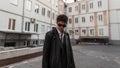 Fashion portrait of a young man hipster in sunglasses with a stylish hairstyle in trendy black leather jacket near buildings on Royalty Free Stock Photo