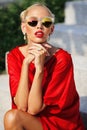 Fashion portrait of young pretty blonde girl in red dress and sunglasses. Beautiful woman model posing outdoor Royalty Free Stock Photo