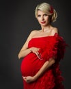 Fashion Pregnant Woman Portrait in Red Dress over Black. Elegant Mother embracing Belly in Luxury Gown with evening Hairstyle. Royalty Free Stock Photo