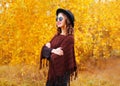 Fashion pretty smiling woman model wearing a black hat sunglasses and knitted poncho over autumn yellow leaves Royalty Free Stock Photo