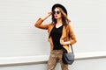 Fashion pretty woman wearing a retro elegant hat, sunglasses, brown jacket and black handbag walking in city over background