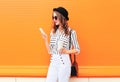 Fashion pretty young woman model using smartphone with coffee cup wearing black hat white pants over colorful orange Royalty Free Stock Photo