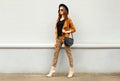 Fashion pretty young woman wearing a retro elegant hat, sunglasses, brown jacket and black handbag walking in city over backg Royalty Free Stock Photo