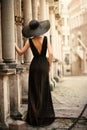 Fashion Woman Model in Hat Back Side View on Old City Street. Elegant Traveler Girl in Black Dress Looking at Antique Architecture